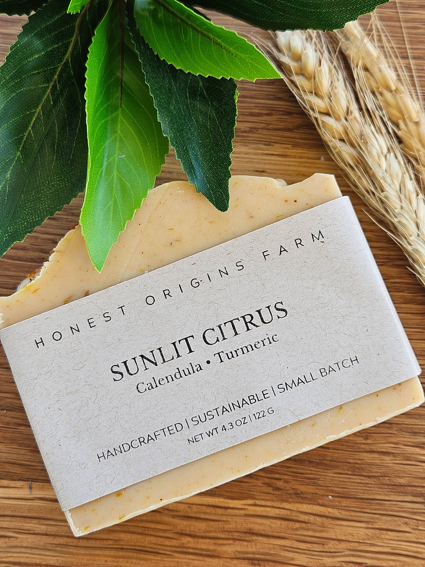 Bar of soap with a label on a wooden surface, surrounded by green leaves and straw.