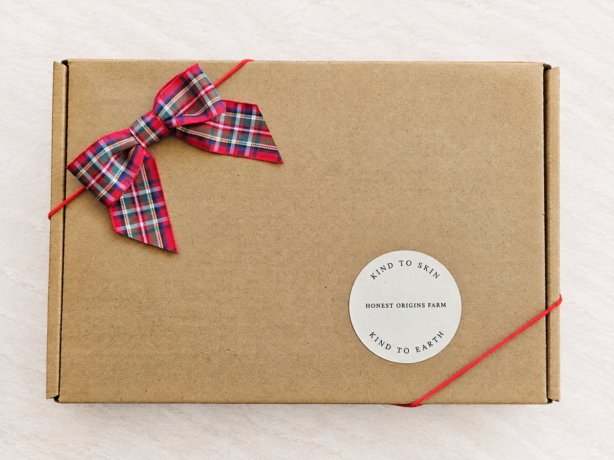 A square brown kraft paper gift box tied with a red string and a red plaid bow, featuring a round white sticker that reads 'KIND TO SKIN HONEST ORIGINS FARM KIND TO EARTH' on a light textured background.