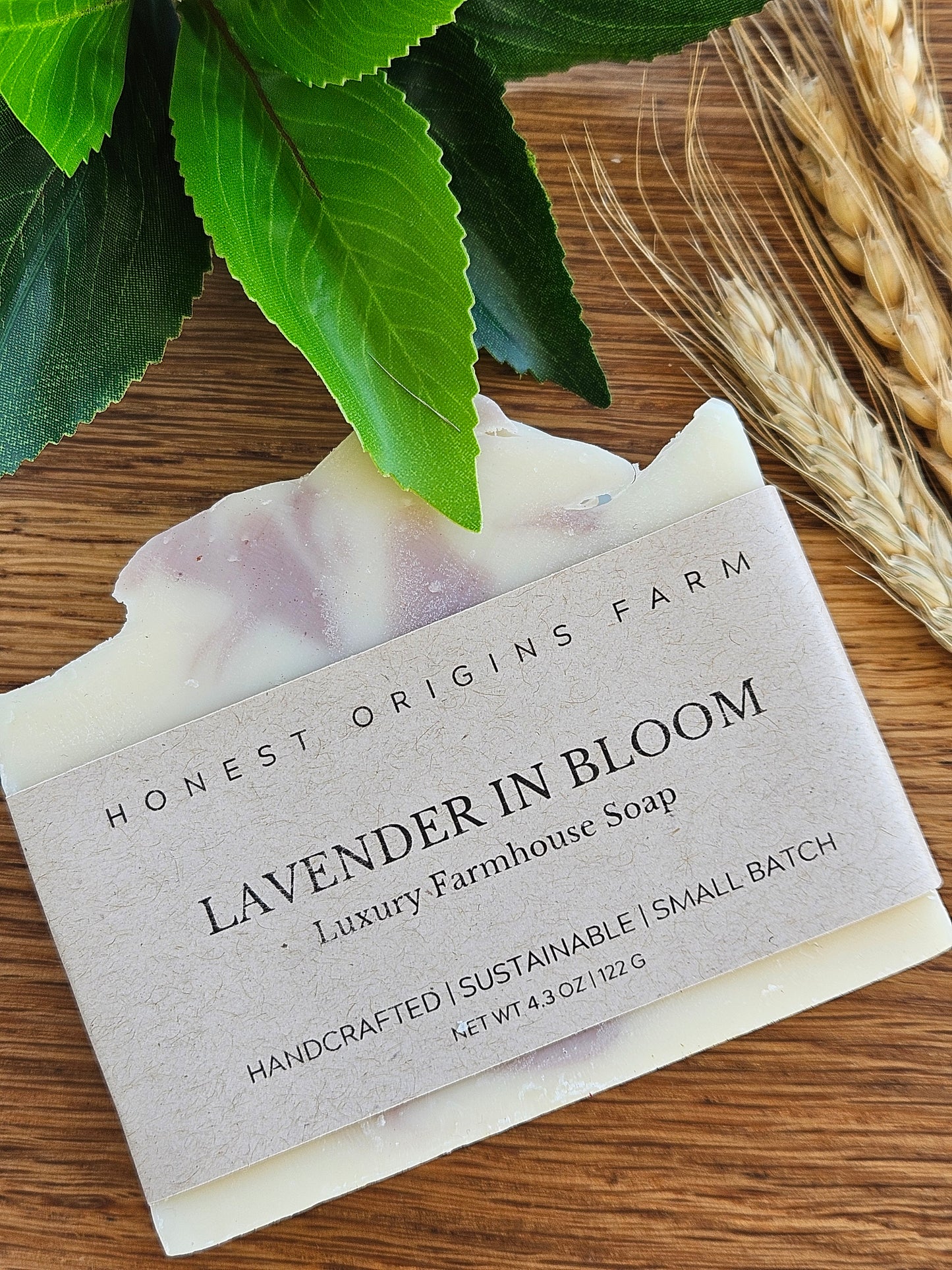 Bar of soap with a label on a wooden surface with green leaves and wheat stalks.