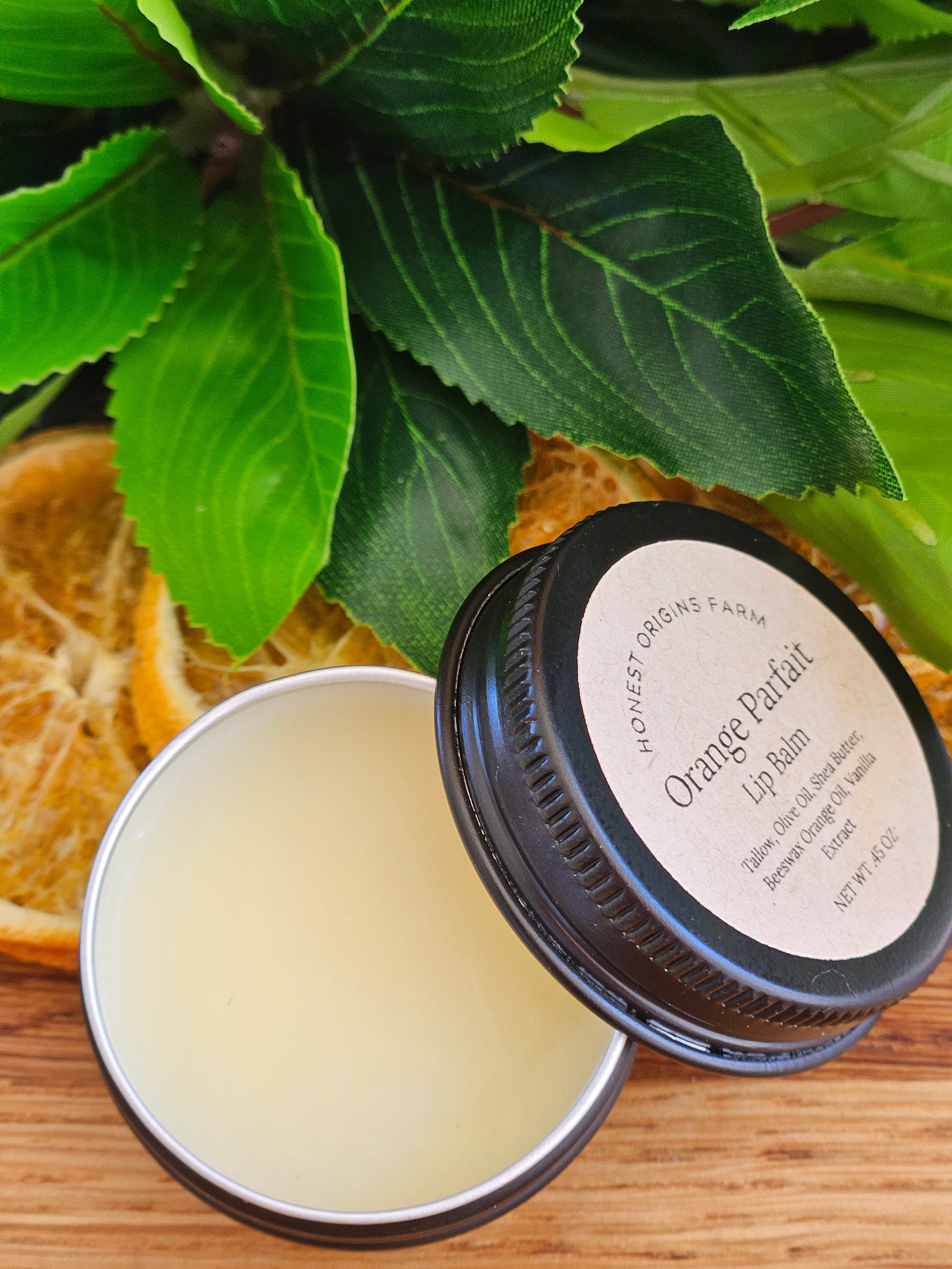 Balm in a tin with a black lid, surrounded by green leaves and oranges on a wooden surface.