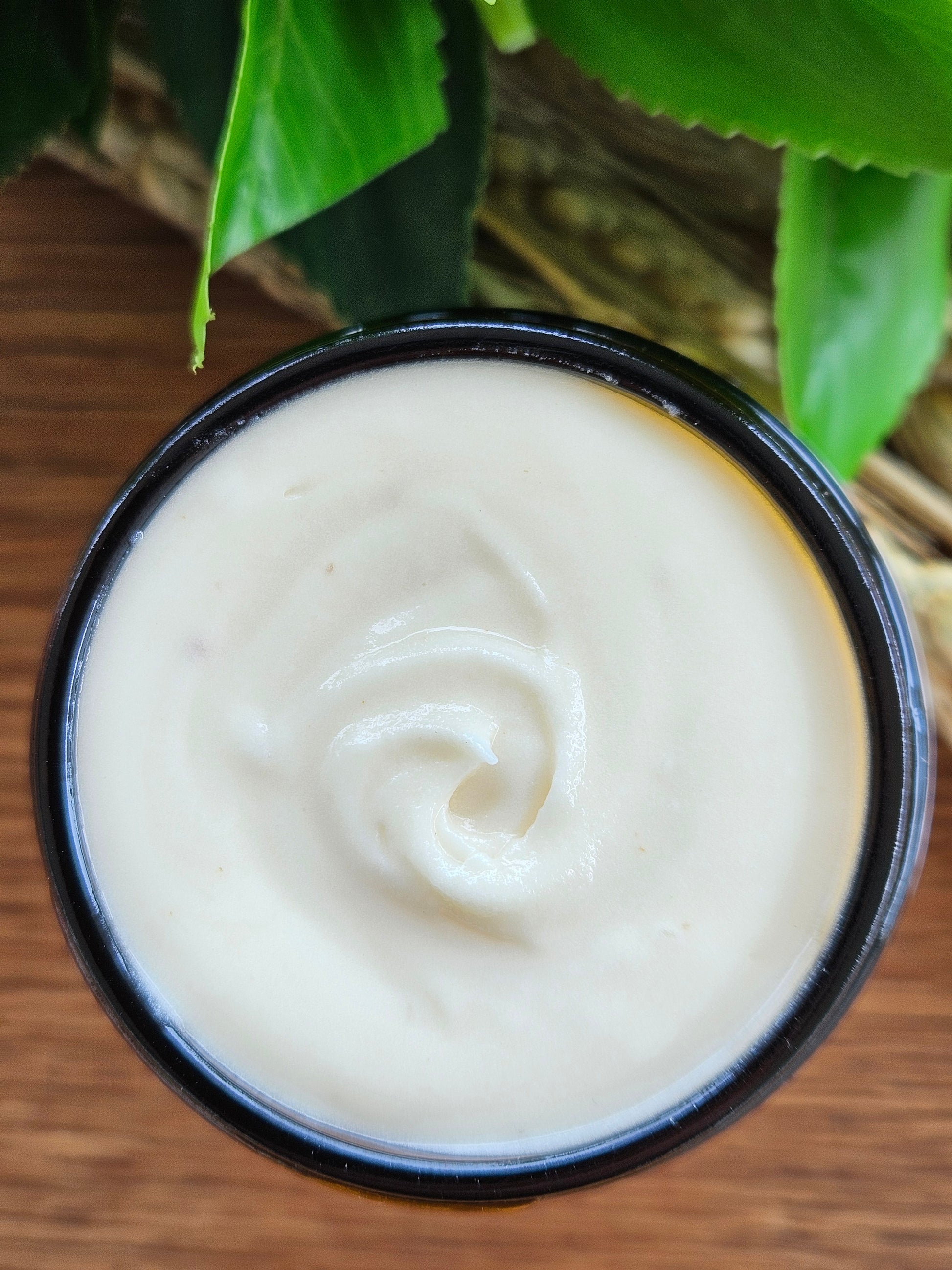 Jar of white cream with a swirl on a wooden surface with green leaves.