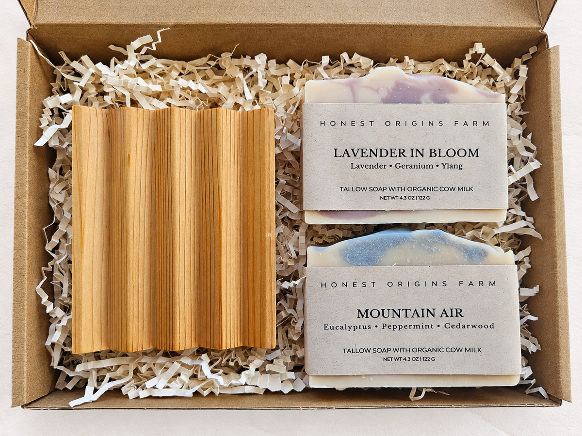 A gift box from Honest Origins Farm containing two tallow soaps, 'Lavender in Bloom' and 'Mountain Air', nestled in white paper shred next to a folded light wood cedar soap dish.